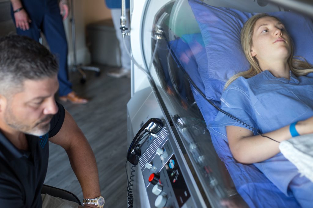 A client sits peacefully in the HBOT at APN Lodge as a technician sits close by outside of the chamber.