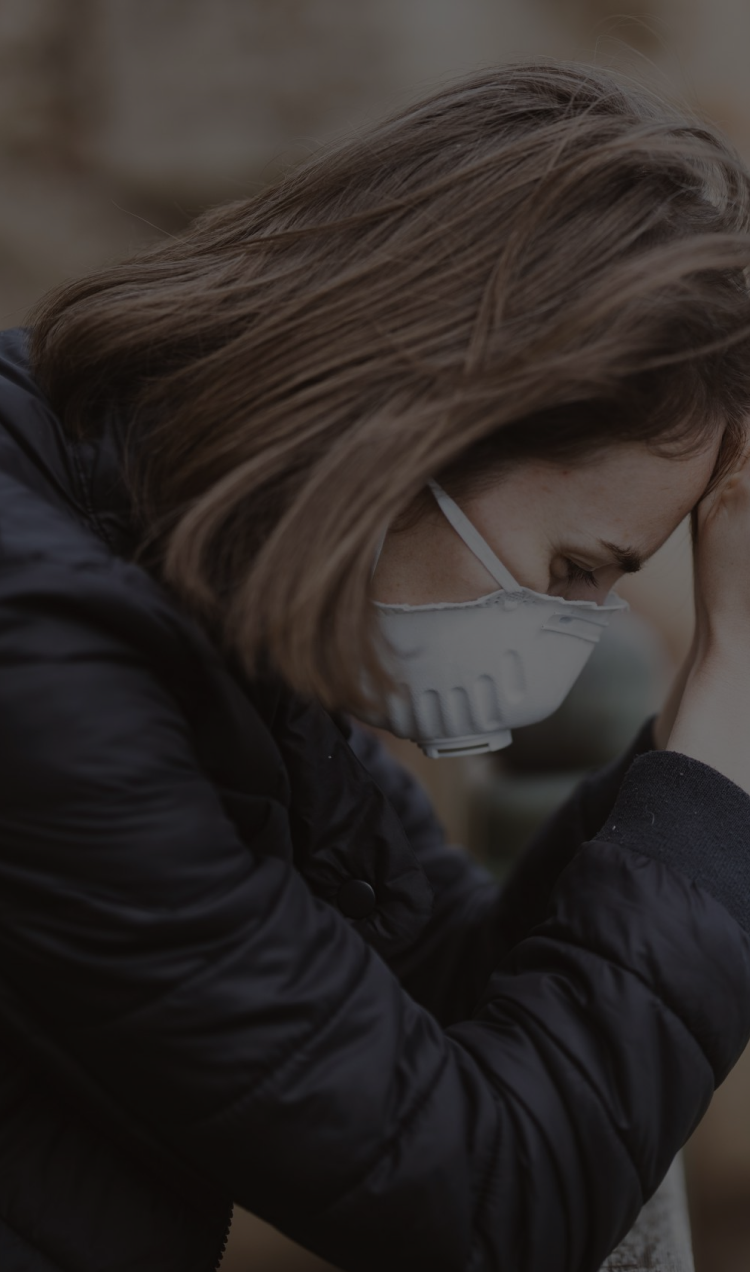 A woman wearing a mask leans with her elbow on a railing in her head in her hands.